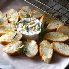 Pieczony camembert z czosnkiem i rozmarynem, otoczony kromkami bagietki skropionymi oliwą, gotowy do zapieczenia.