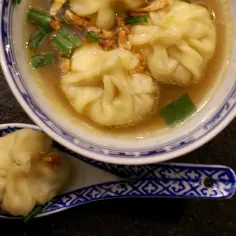 A miseczce z niebiesko-białym wzorem znajduje się aromatyczna zupa wonton z zieloną cebulką i chrupiącymi dodatkami. Na łyżce leży jeden pierożek wonton.