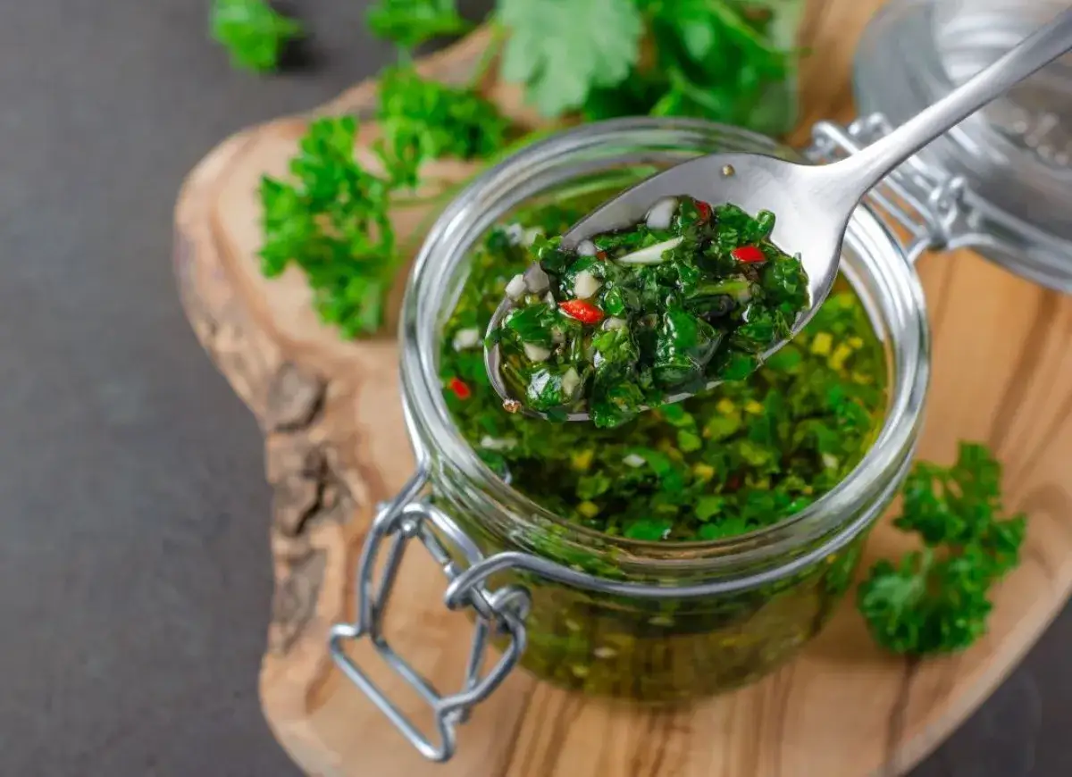 Łyżka pełna aromatycznego sosu chimichurri, z widocznymi kawałkami ziół i papryczki chili, nad słoikiem pełnym tego zielonego skarbu.