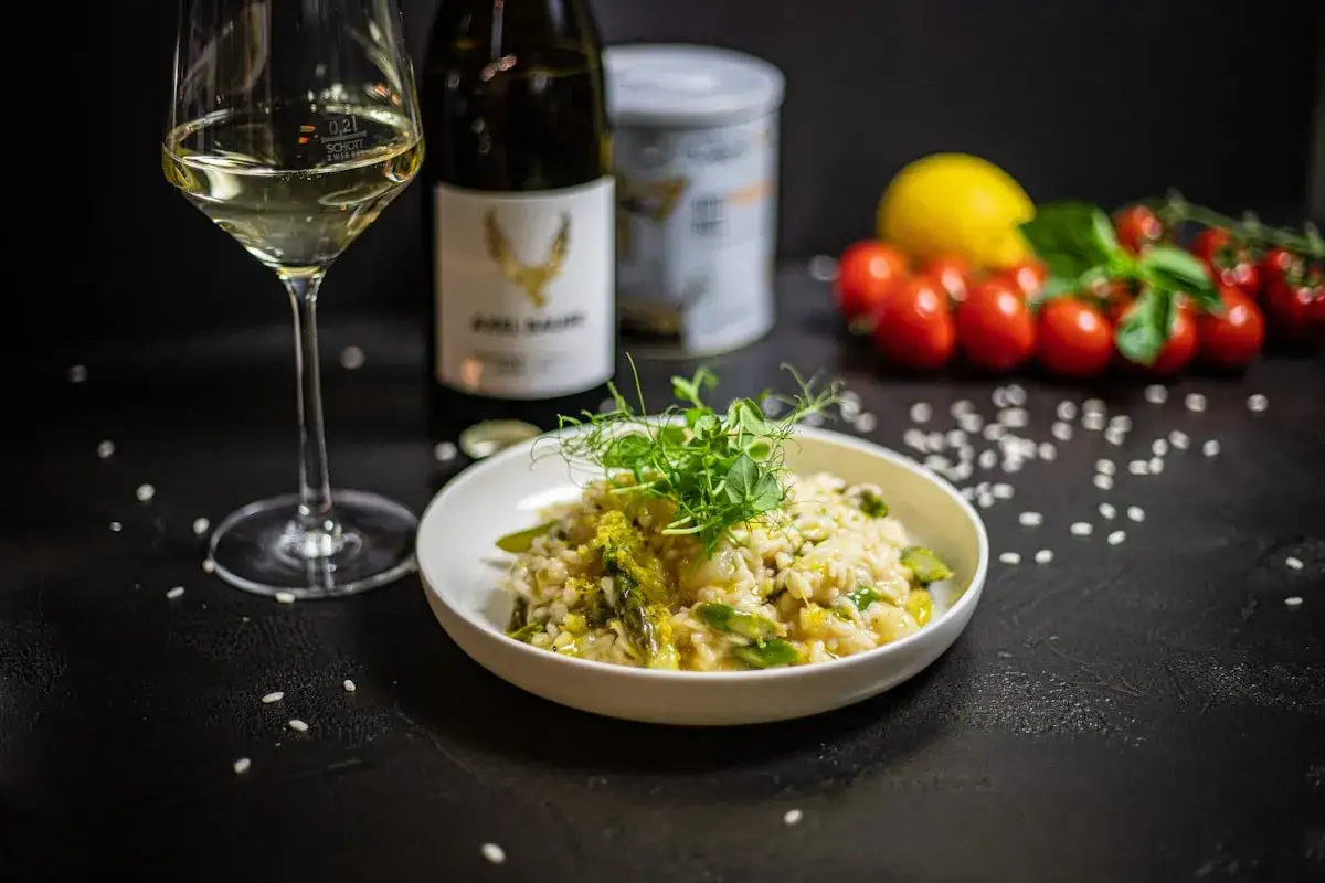 Klasyczne risotto z kurczakiem, podane z zielonymi warzywami i kiełkami, obok kieliszka białego wina i butelki.