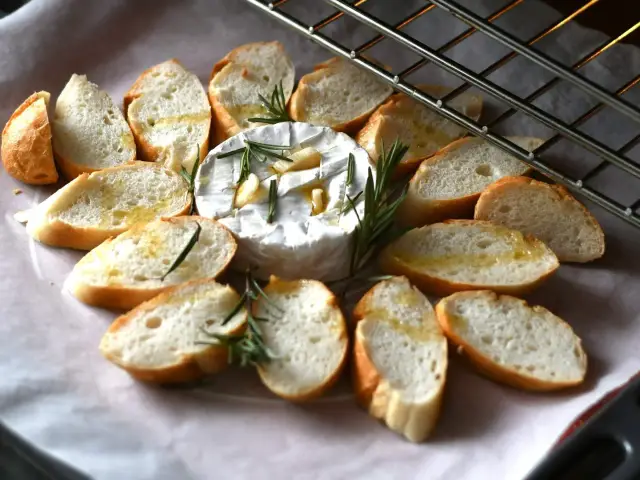 Pieczony camembert z czosnkiem i rozmarynem, otoczony kromkami bagietki skropionymi oliwą, gotowy do zapieczenia.